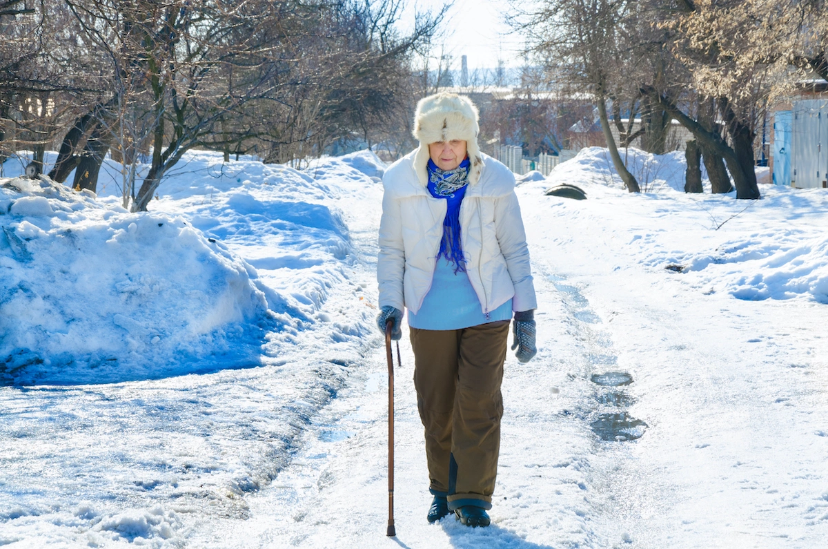 Senior female in white fur trapper hat walking carefully in snow with cane