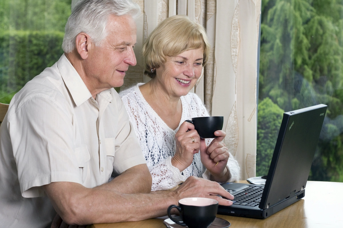 Senior couple including male and female seated at laptop computer with coffee cups appearing pleased with telemedicine session