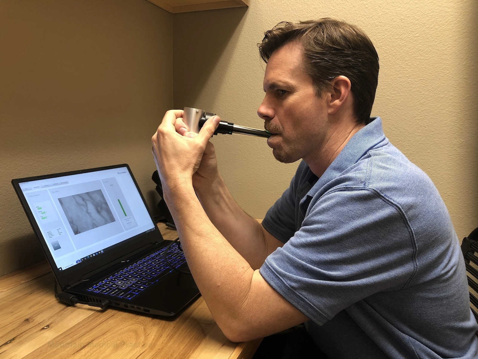 GlycoCheck video camera positioned under the tongue of a young adult male seated at laptop computer screen capturing microscopic video image of red blood cells moving inside capillaries