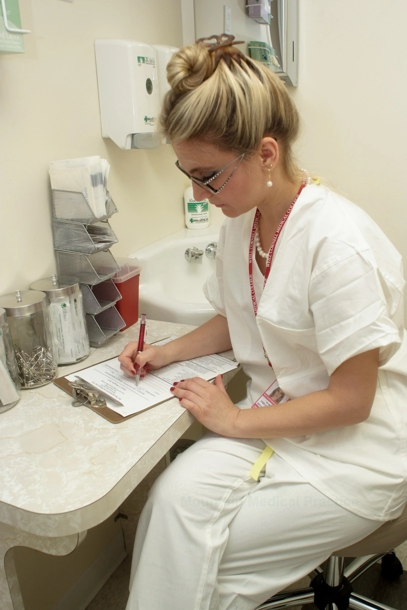 Dr. Maria Pirraglia in white scrubs seated at small exam room desk writing with pen on medical form