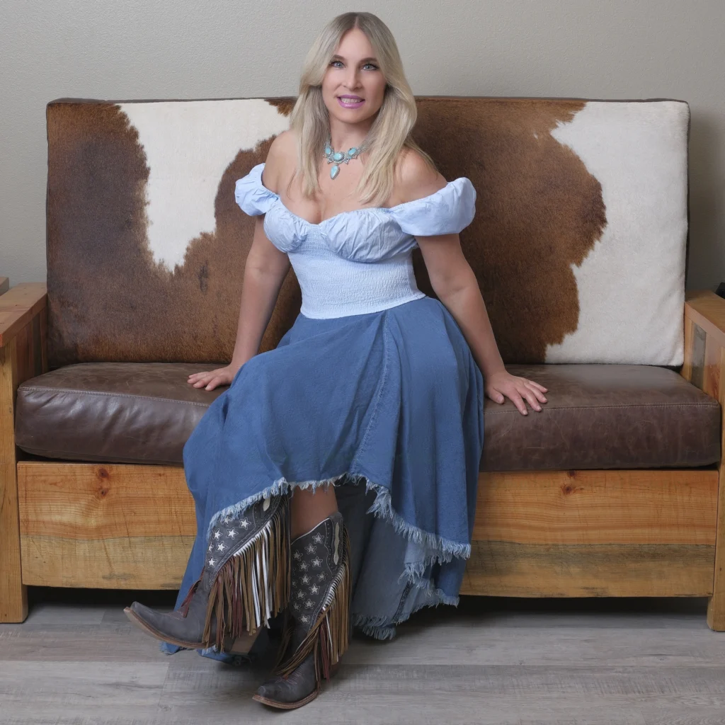 Dr. Maria Pirraglia in light blue peasant blouse, denim maxi skirt, fringe cowgirl boots, and turquoise necklace seated on rustic cowhide couch in waiting room