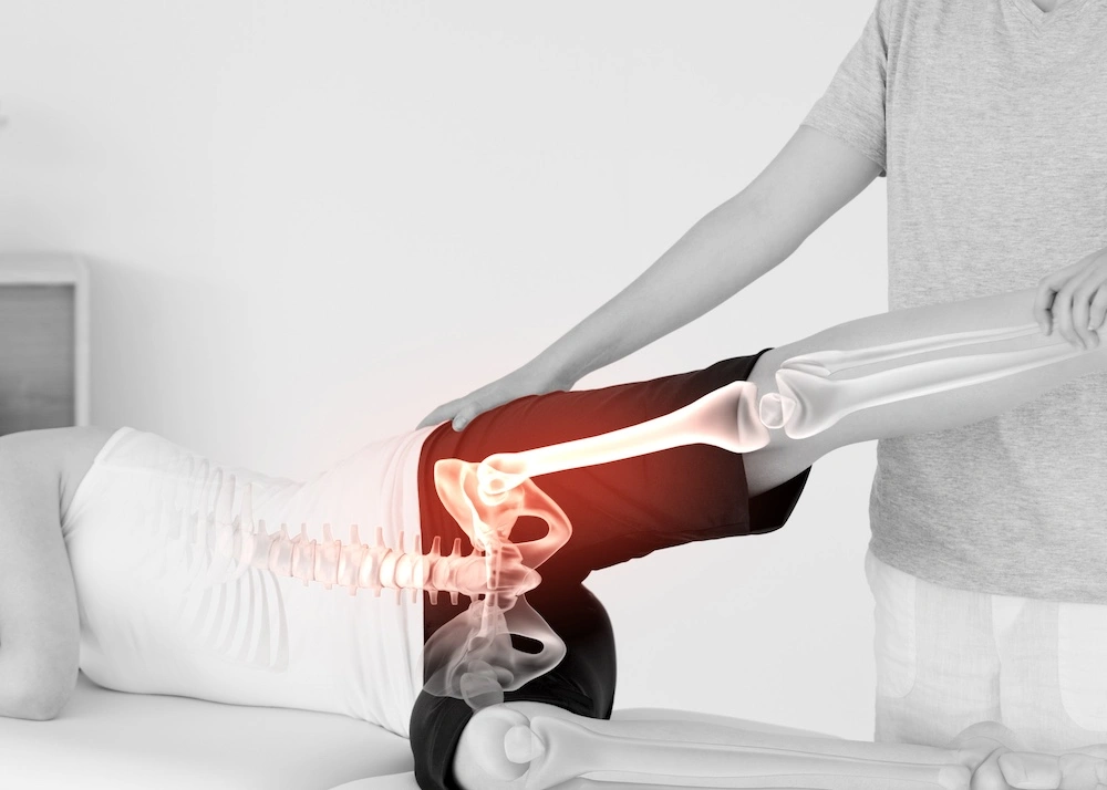 Female lying sideways on exam table with inset hologram of painful hip joint receiving assistance from female physical therapist