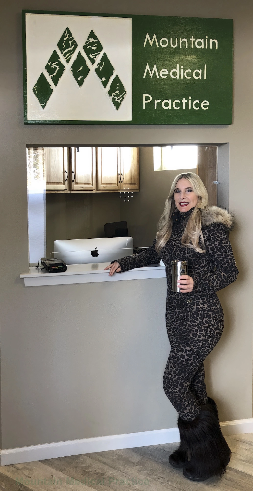Dr. Maria Pirraglia cheerful in Bogner ski apparel leopard onesie with silver beverage tumbler in hand standing at front reception area in alpine office setting under Mountain Medical Practice signage