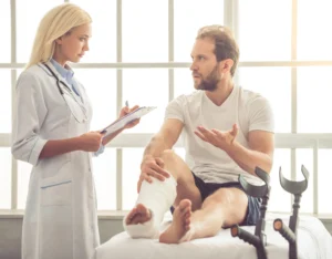 Female physiatrist standing beside exam table assisting male patient wearing orthopedic cast on injured right leg with Lofstrand forearm crutches